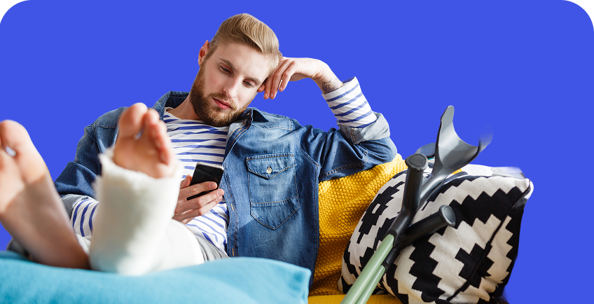 Vyras su sulaužyta ir sugipsuota koja sėdi ant sofos ir naršo mobiliajame telefone