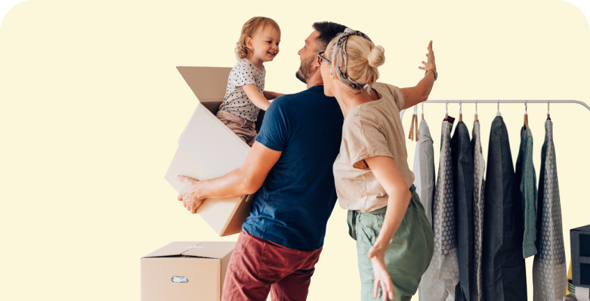 Tėtis laiko didelę dėžę, kurioje stovi nedidelis vaikas, o šalia stovi mama ištiesusi rankas, visi šypsosi.