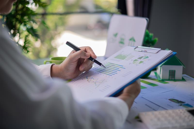 Žmogus rankose laiko tušinuką ir susegtus popierinius grafikus, ir juos nagrinėja.