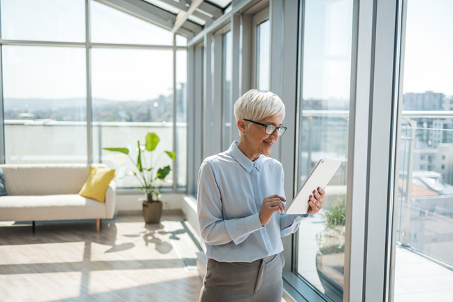 Verslininkė šviesiame bute su dideliais langais naudojasi planšete.